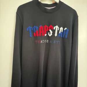 Svart  sweatshirt med brodyr - Svart sweatshirt med broderad logga i blått, rött och vitt framtill. Tröjan har rund halsringning och långa ärmar. Texten 'IT'S A SECRET' är broderad under loggan i matchande färger. Perfekt för en streetwear-look.  Köpt på 700 kr knappt använd