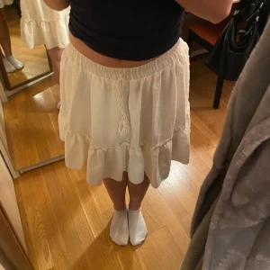 Vit volangkjol med snörning - Supersöt vit kortkjol med volangkant och snörning i midjan. Kjolen har en luftig och flowy passform, perfekt för varma dagar. Materialet känns mjukt och bekvämt mot huden. 