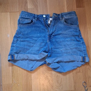 Blå högmidjade jeansshorts - Snygga blå jeansshorts med hög midja och klassisk femficksdesign. Shortsen har uppvikta ben och stängs med knapp och dragkedja. Materialet är 83% bomull, 16% polyester och 1% elastan vilket gör dem både bekväma och slitstarka. Perfekta för varma dagar.