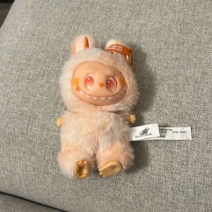 Labubu  - Bilderna visar en mjukisdocka från POP MART, serien THE MONSTERS. Dockan har ett lurvigt, ljusrosa yttre och ett glatt, fantasifullt ansikte med stora ögon och ett brett leende. Perfekt som samlarobjekt eller rolig dekoration för unga och unga vuxna som gillar unika figurer.