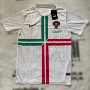 Portugal fotbollströja Nike 2012 - Portugal fotbollströja från Nike. Vit bas med gröna och röda korsade linjer på framsidan, broderat landslagsemblem och Dri-FIT-material. Kortärmad modell, perfekt för dig som älskar fotboll och vill sticka ut med en ikonisk tröja.