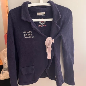 Mörkblå cardigan jacka Odd Molly - Mysig mörkblå cardigan jacka från Odd Molly med broderad text och rosdetalj på ryggen. Jackan har lång ärm, krage och knytband i ljusrosa framtill. Perfekt för dig som gillar unika detaljer och skön passform.