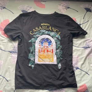 Svart Avenida Casablanca t-shirt - Svart t-shirt med färgglad tryckt grafik på både bröstet och ryggen. Motivet föreställer en byggnad med texten 'Avenida Casablanca' i gult och gröna blad runt fönstret. Klassisk passform och rund hals. Perfekt för dig som gillar unika prints och streetstyle.