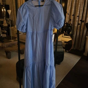 Ljusblå långklänning med puffärm - Superfin ljusblå långklänning med puffiga kortärmar och rund halsringning. Klänningen har en luftig passform och volangdetaljer nertill för en extra flowy känsla. Perfekt för dig som gillar en romantisk och trendig stil.