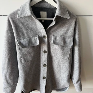 Grå jacka från H&M  - Snygg grå jacka från H&M  med klassisk krage, stora bröstfickor och knappar framtill. Modellen är lite oversized och har slits i sidorna. 