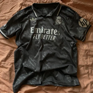 Real Madrid Mbappé fotbollströja - Snygg svart Real Madrid fotbollströja från Adidas med Mbappé och nummer 9 på ryggen. Tröjan har korta ärmar, klassisk krage och silvriga detaljer. Tillverkad i lätt och ventilerande material med diskret mönster och tryckta loggor på bröst och ärm.