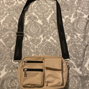 Beige axelväska med svarta detaljer - Snygg beige axelväska med bred svart justerbar rem. Väskan har flera praktiska fack framtill med dragkedja och lock, samt ett rymligt huvudfack. Perfekt för att hålla koll på dina grejer när du är på språng. Materialet är lätt och smidigt syntet.