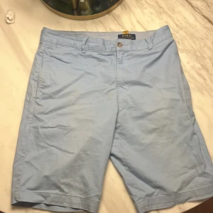 Ljusblå shorts från Polo Ralph Lauren - Snygga ljusblå shorts från Polo Ralph Lauren med klassisk design och raka ben. De har fickor fram och bak med knappstängning och bälteshällor. Materialet är mjuk bomull som känns chill och fräscht under varma dagar. Perfekt för dig som gillar stilren och enkel look. Storleken är 18 som passar ungefär xs.                                                  Mvh Teo