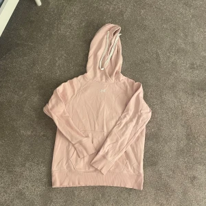 Ljusrosa hoodie från Under Armour - Säljer en ljusrosa hoodie från Under Armour med vit logga på bröstet. Tröjan har huva med snörning och en stor magficka. Perfekt för dig som gillar en avslappnad och sportig stil.
