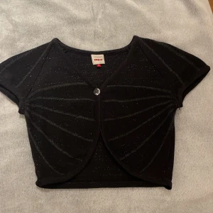 Glittrig svart bolerotopp från ONLY❤️ - Säljer en svart glittrig bolerotopp från ONLY i storlek M. Toppen är croppad med korta ärmar, v-ringning och en snygg knapp framtill. Materialet är stickat med ränder och lite glitter! Älskar denna egentligen, så söt, men den kommer tyvärr inte till användning.❤️Har knappt använt den.