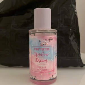 Unibow Dream Eau de Toilette - Söt parfymflaska med pastellrosa och blå etikett, dekorerad med enhörningsmotiv och regnbågar. Ljuslila plastlock och genomskinlig glasflaska. Doften är framtagen för henne och har en lekfull, drömmig design som sticker ut på hyllan.