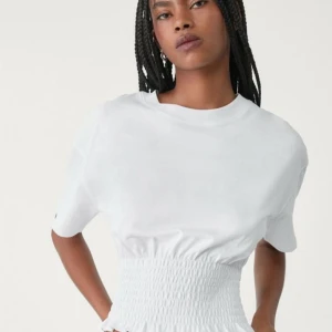 Vit croppad t-shirt från Strteen - Säljer en vit croppad t-shirt från Strteen med smockad midja och volangkant nedtill. T-shirten har korta ärmar och är gjord i mjuk bomull, perfekt för en chill och trendig look. Enkel att matcha med både jeans och kjol.