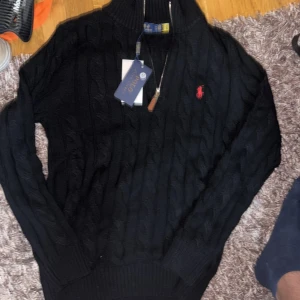 Svart stickad tröja Polo Ralph Lauren - Svart kabelstickad tröja från Polo Ralph Lauren med halv dragkedja och klassisk röd logga broderad på bröstet. Tröjan har hög krage, långa ärmar och ribbade muddar. Perfekt för dig som gillar en stilren och tidlös look.