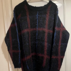 Mörk rutig stickad tröja - Säljer en oversized stickad tröja med rutig design i svart, vinrött och blått. Tröjan har en fluffig, grovstickad känsla och rund hals. Perfekt för dig som gillar en chill och mysig vibe med lite edge.