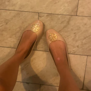 Rosa ballerinaskor från Tory Burch - Snygga rosa ballerinaskor från Tory Burch med rund tå och låg klack. Skorna har en glansig finish och är dekorerade med det klassiska guldiga Tory Burch-emblemet framtill. Perfekta för dig som gillar stilrena och eleganta detaljer.