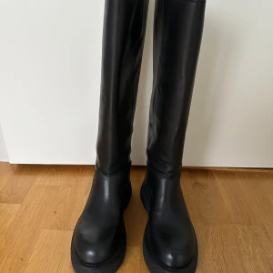 Svarta höga boots från Arket - Snygga svarta boots från Arket i kromfritt skinn med chunky platt sula. Högt skaft och minimalistisk design, perfekta till höst- och vinterlooken. Tillverkade i Portugal och riktigt trendiga till både jeans och kjol.