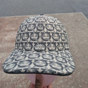 Salvatore Ferragamo mönstrad keps - Snygg keps från Salvatore Ferragamo med grått och beige mönster över hela kepsen. Justerbar rem baktill med metallspänne och svart läderdetalj på sidan. Tillverkad i bomull och polyester, perfekt för dig som gillar exklusiva accessoarer.