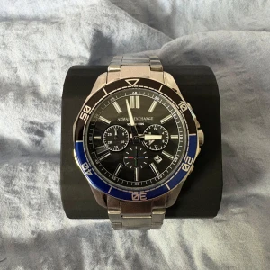 Armani Exchange klocka med blå detalj - Stilren klocka från Armani Exchange med robust länkarmband i silverfärgat metall. Urtavlan är svart med blå och silvriga detaljer, tre mindre urtavlor och tydliga index. Klockan har en sportig vibe och levereras i originalbox med matchande AX-halsband.