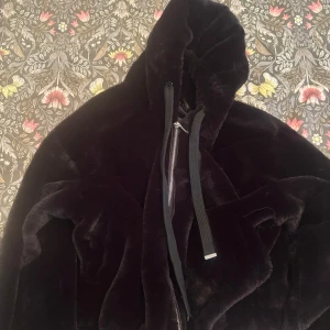 Svart pälsjacka från Jennyfer - Mysig svart pälsjacka från Don't Call Me Jennyfer med huva och dragkedja framtill. Jackan har breda snören vid huvan och är perfekt för kalla dagar när du vill vara både varm och snygg. Fodrad insida för extra komfort.