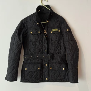 Svart quiltad jacka från Barbour strl.34 - International Polarquilt från Barbour med bälte i midjan, guldiga knappar och dragkedja. Klassisk look med flera fickor och Barbour International-logga på bröstet. Perfekt för dig som gillar stilren och funktionell design. Sparsamt använd. 