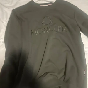 Grön Moncler sweatshirt med broderad logga - Snygg grå sweatshirt från Moncler med broderad logga och emblem framtill. Tröjan har rund halsringning och långa ärmar, perfekt för en clean och stilren look. Ribbstickade muddar vid ärmslut och nederkant för extra komfort.