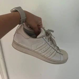 Vita Adidas Superstar sneakers - Klassiska vita Adidas Superstar sneakers med ikoniska tre ränder på sidan och snörning. Skorna har rund tå, platt sula och är tillverkade i skinn. Perfekta för dig som gillar en clean och tidlös streetstil.