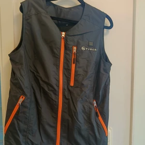 Svart värmeväst från Pumori XS - Svart värmeväst från Pumori i syntetmaterial med orange dragkedjor och flera fickor. Västen har inbyggd värmefunktion som drivs av medföljande powerbank. Perfekt för kalla dagar och utomhusaktiviteter. Snygg, sportig look med diskret logga på bröstet.