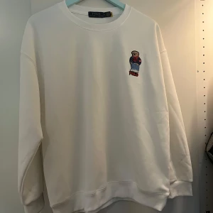 Vit sweatshirt Polo Ralph Lauren M - Vit sweatshirt från Polo Ralph Lauren med broderad nalle på bröstet. Klassisk rund halsringning och avslappnad passform. Perfekt för dig som gillar stilrena plagg med en lekfull detalj. Materialet är mjuk bomull.
