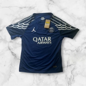 Blå PSG fotbollströja med vita detaljer - Snygg blå PSG fotbollströja med vita vingmönster på axlarna och Qatar Airways-tryck på bröstet. Tröjan har korta ärmar, rund hals och är tillverkad i ett lätt och ventilerande material. Perfekt för fotbollsträning eller som supporterplagg.