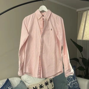 Rosa och vit randig skjorta från Polo Ralph Lauren - Säljer en klassisk rosa och vit randig skjorta från Polo Ralph Lauren i custom fit. Skjortan har lång ärm, knappar framtill och broderad logga på bröstet. Perfekt för dig som gillar stilrena och tidlösa plagg.