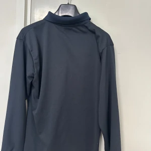 Svart långärmad pikétröja från Polo Ralph Lauren - Klassisk svart långärmad pikétröja från Polo Ralph Lauren med vit broderad logga på bröstet. Tröjan har krage och knappar framtill, perfekt för en stilren look.