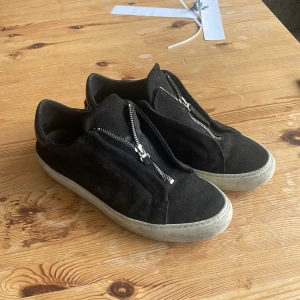 Axel arigato skor  - Svarta sneakers i mocka med vit platt sula och dragkedja framtill istället för snörning. Skorna har en rund tå och en stilren design som passar till många outfits.                                                                                               Dust bag medföljer