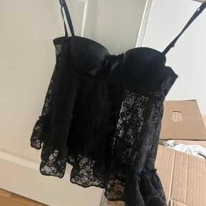 Svart spetsig korsettopp - Säljer en svart korsettopp med tunna axelband och vackra spetsdetaljer. Toppen har bygel och lätt volang nertill för en extra feminin touch. Perfekt att styla till olika outfits!