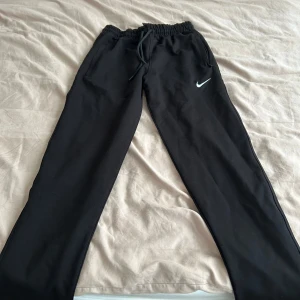 Svarta träningsbyxor från Nike - Svarta träningsbyxor från Nike med vit logga på låret. Byxorna har dragsko i midjan och praktiska fickor med dragkedja. Perfekta för gymmet eller löprundan, med en bekväm och normal passform.