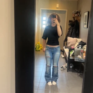 Ljusblå slitna bootcut jeans - Säljer ett par ljusblå jeans med bootcut och slitna detaljer vid knäna. Jeansen har låg midja och klassisk femficksdesign. Perfekta för en avslappnad och trendig look.