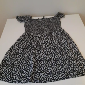 Blommig svart klänning med kort ärm - Söt svart klänning med vitt blommönster och korta ärmar. Klänningen har smock upptill och en lätt utställd kjol. Perfekt för varma dagar och enkel att styla med sneakers eller sandaler.