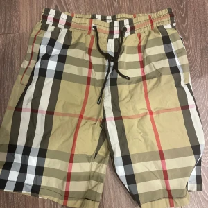 Burberry shorts  - Snygga beige shorts med klassiskt rutigt mönster i svart, vitt och rött. De har elastisk midja med svart snörning och sidofickor. Perfekta för sommaren och har en avslappnad passform.