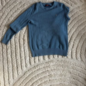 Blå stickad tröja från Morris - Säljer en blå stickad tröja från Morris med rund halsringning och diskret broderad logga på bröstet. Tröjan har lång ärm och ribbade muddar vid ärmslut och nederkant. Perfekt för lager-på-lager och enkel att matcha.