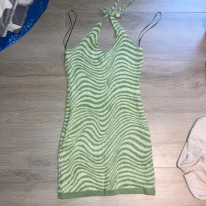 Grön stickad halterneckklänning med vågmönster - Säljer en grön stickad halterneckklänning med vågigt mönster. Klänningen har smala axelband och knyts i nacken. Perfekt för dig som gillar unika mönster och vill sticka ut lite extra.