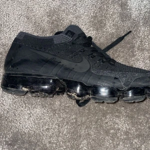 Svarta Nike Vapormax sneakers - Säljer ett par helsvarta Nike Vapormax sneakers med stickad ovandel och tydlig Nike-logga på sidan. Skorna har snörning och den klassiska genomskinliga bubbliga sulan som ger en futuristisk look. Perfekta för dig som gillar streetwear och sportig stil.