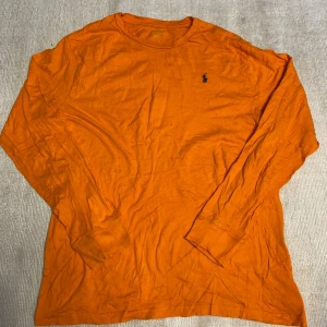 Orange långärmad tröja från Polo Ralph Lauren - Säljer en orange långärmad tröja från Polo Ralph Lauren med klassisk broderad logga på bröstet. Tröjan har rund halsringning och är tillverkad i mjuk bomull, perfekt för en avslappnad stil.