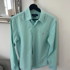 Skjorta från Polo Ralph Lauren - Säljer en  långärmad skjorta från Polo Ralph Lauren i slim fit-modell. Skjortan har klassisk krage, knäppning framtill och en rosa broderad logga på bröstet. Perfekt för dig som gillar stilrena och tidlösa plagg. 