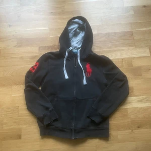 Ralph lauren hoodie  - Svart hoodie med dragkedja från Polo Ralph Lauren. Stor röd broderad logga på bröstet och siffra på ärmen. Grå insida på huvan och vita snören. Perfekt för en avslappnad stil.