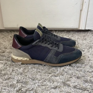 Valentino Garavani sneakers i blått och grått - Säljer ett par Valentino Rockrunner sneakers i blått, beige och vinrött. Skorna har klassisk snörning, rund tå och detaljer i mocka och skinn. Storlek 45 men passar 46, skriv för mer bilder eller mått! Dustbag kommer med!