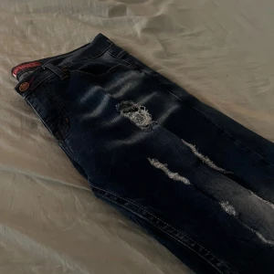 Mörkblå slitna jeans - Säljer ett par mörkblå jeans med slitningar och snygga dragkedjor på bakfickan. Jeansen har klassisk femficksdesign och en läderpatch bak i midjan. Perfekta för dig som gillar en lite tuffare stil.