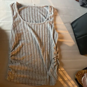 Grå ribbad linnetopp - Säljer en grå ribbad linnetopp med rynkade detaljer på sidan. Toppen är ärmlös och har en rund halsringning. Perfekt för en avslappnad och stilren look.