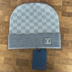 Grå rutig mössa från Louis Vuitton - Snygg grå mössa från Louis Vuitton med klassiskt rutigt mönster och vikt kant. LV-logga framtill på kanten. Perfekt accessoar för kyliga dagar.