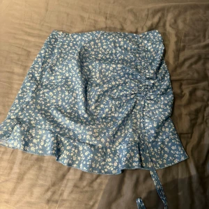 Blå blommig omlottkjol med volang - Supersöt blå kjol med vitt blommönster och volangkant. Kjolen har omlottmodell med knytband i sidan och en lätt rynkad detalj framtill. Perfekt för en fräsch och somrig look!