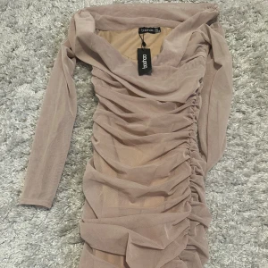 Beige omlottklänning från Boohoo - Säljer en beige långärmad klänning från Boohoo med snygg omlottdesign och rynkade detaljer längs sidorna. Klänningen har en figurnära passform och är tillverkad i ett tunt, mesh-liknande material. Aldrig använt. 