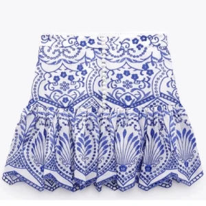 Vit och blå volangkjol från Zara - Supersöt kort kjol i vitt med blått broderat mönster och volang nertill. Kjolen är perfekt för sommarens alla tillfällen. En liten fläck som man ser på bild 3 annars super fin!!! 